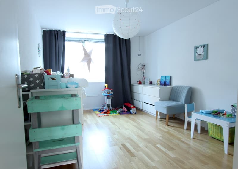Brightly lit children's room with wooden flooring, large window with dark curtains, a white hanging lamp, a grey upholstered chair, a small white table with a blue surface, a green wooden chair, a white cabinet, and various toys scattered around.