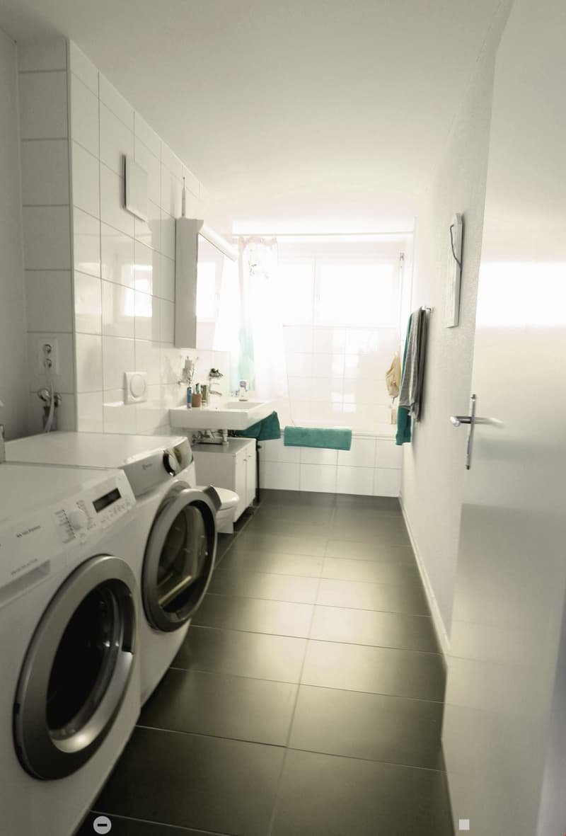 Modern bathroom with a washing machine, sink, mirror, and towels. The space is well-lit with natural light from a window and features tiled walls and a tiled floor.