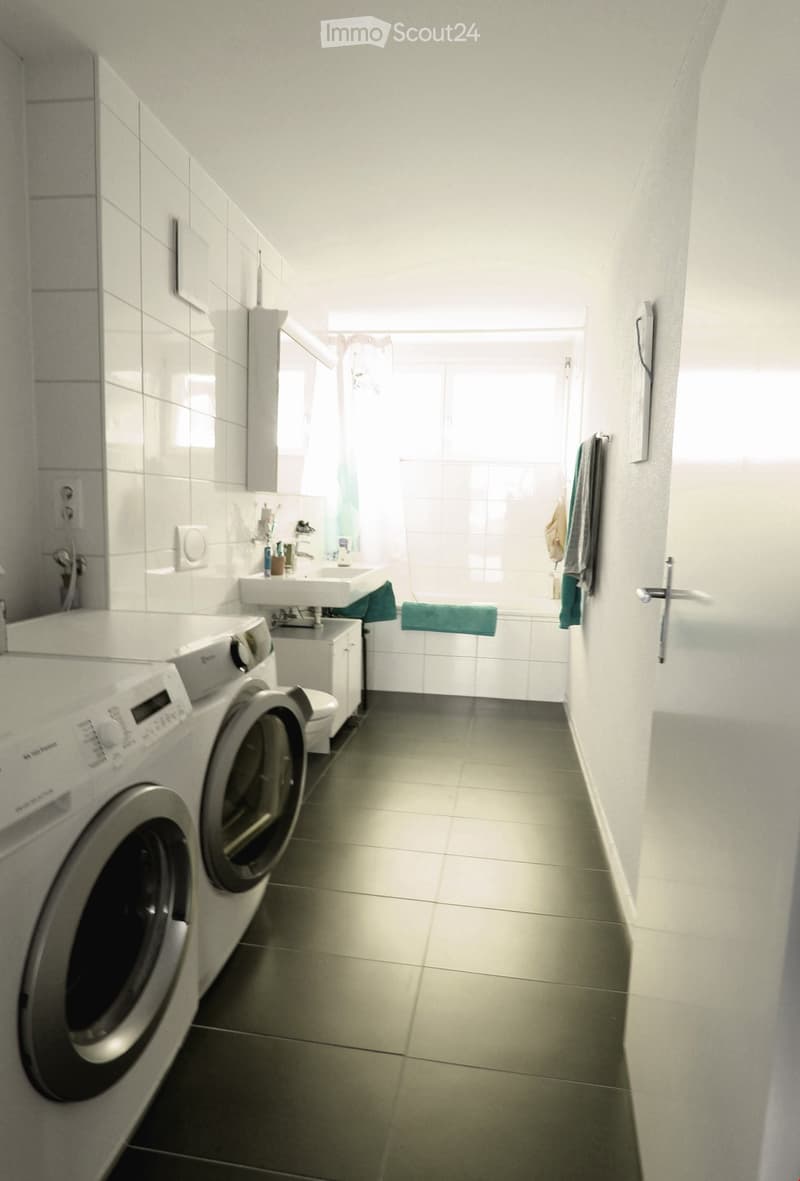 Modern bathroom with a washing machine, sink, mirror, and towels. The space is well-lit with natural light from a window and features tiled walls and a tiled floor.