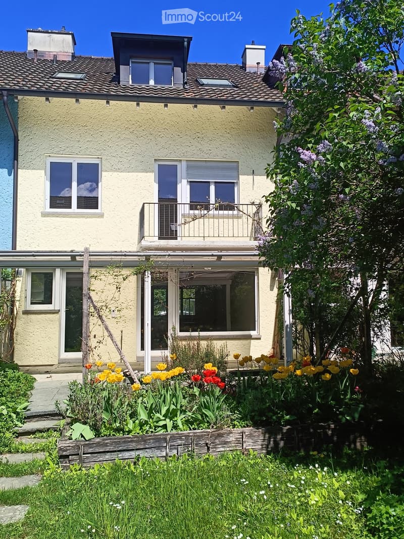 Two-story house with yellow exterior, multiple windows, and a balcony. The garden features colorful tulips and a wooden planter box.