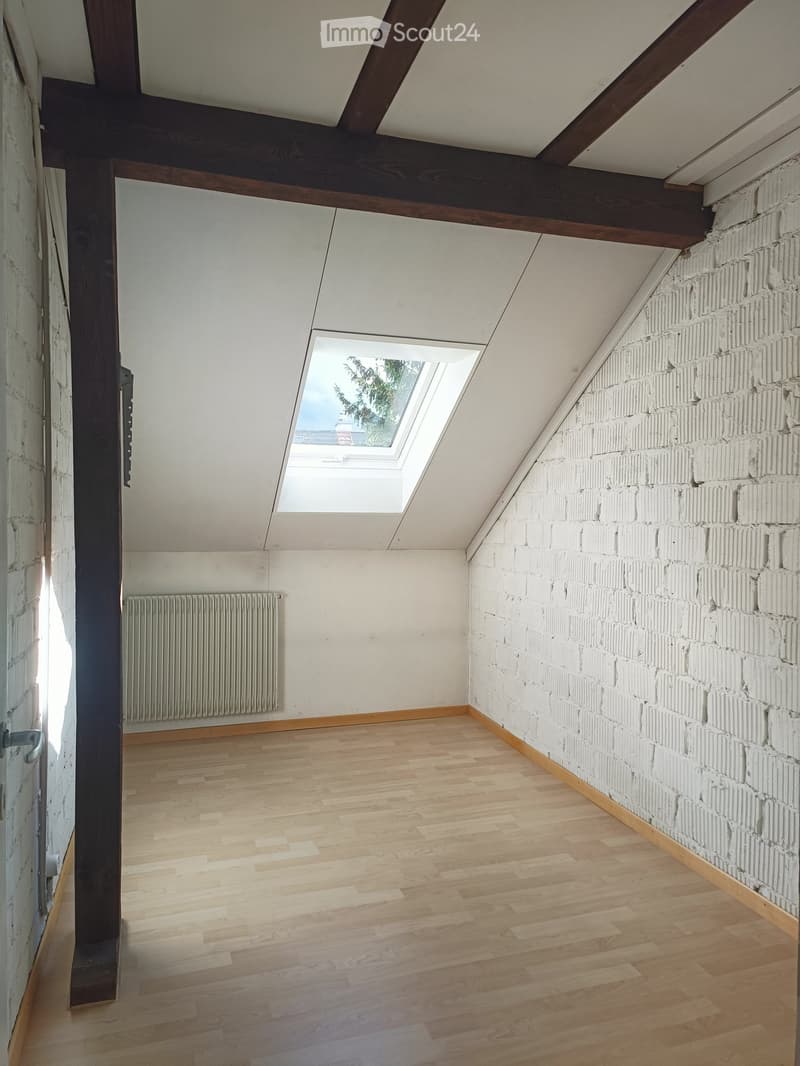 Empty attic room with wooden flooring, exposed wooden beams, white brick wall, skylight window, and radiator.