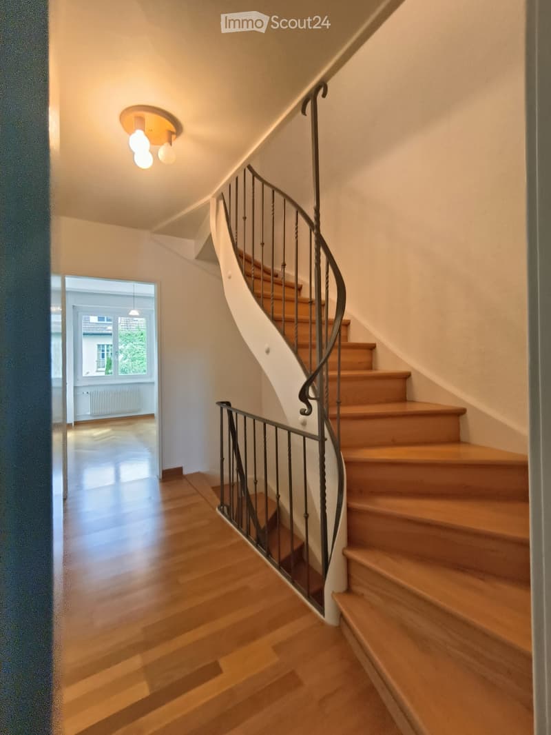 Spiral staircase with wooden steps and metal railing, leading to a bright room with large windows and wooden flooring.