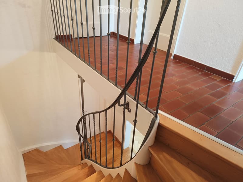 Spiral staircase with wooden steps and metal railing, leading to an upper level with terracotta tiles.