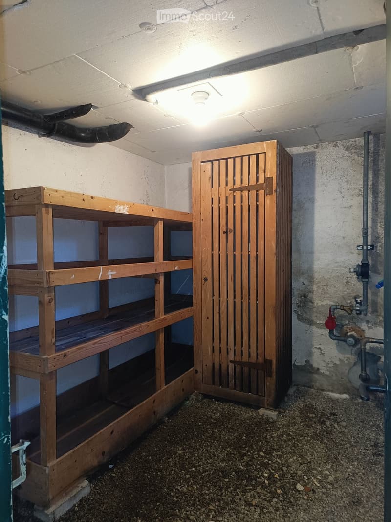 Empty storage room with wooden shelving units and a wooden door, concrete walls, and a gravel floor.