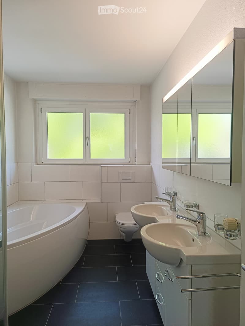 Modern bathroom with a bathtub, toilet, double sink, and large mirror. Dark tiled floor and white tiled walls.