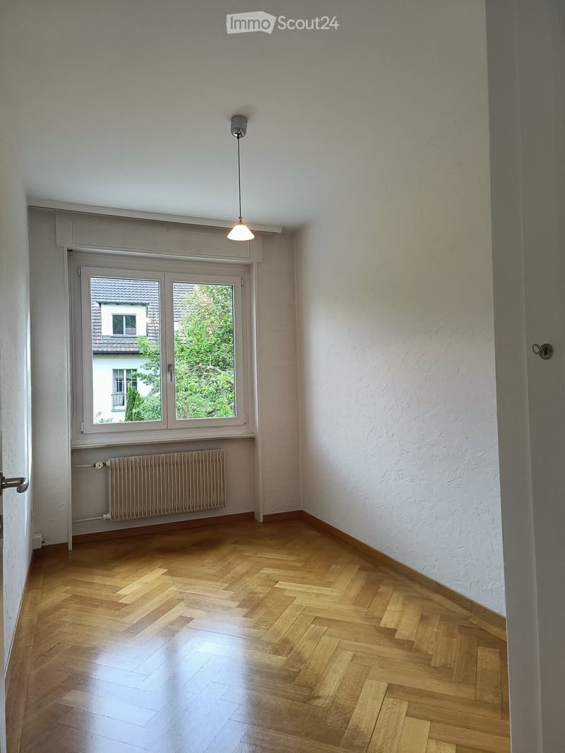 Empty room with wooden herringbone flooring, white walls, a window with a view of a neighboring building and greenery, a radiator below the window, and a single pendant light fixture hanging from the ceiling.