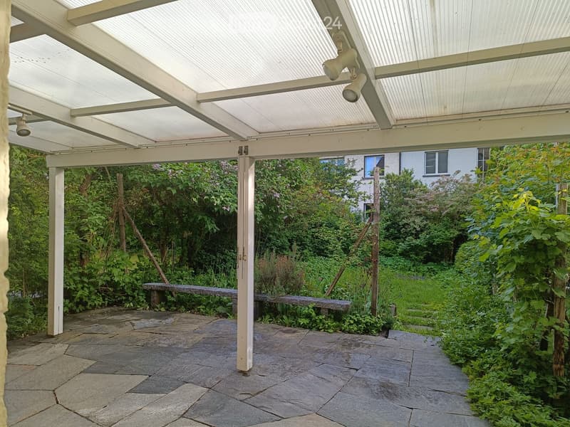 A covered outdoor area with a stone patio, surrounded by lush greenery and benches, with a white-roofed structure and visible house in the background.