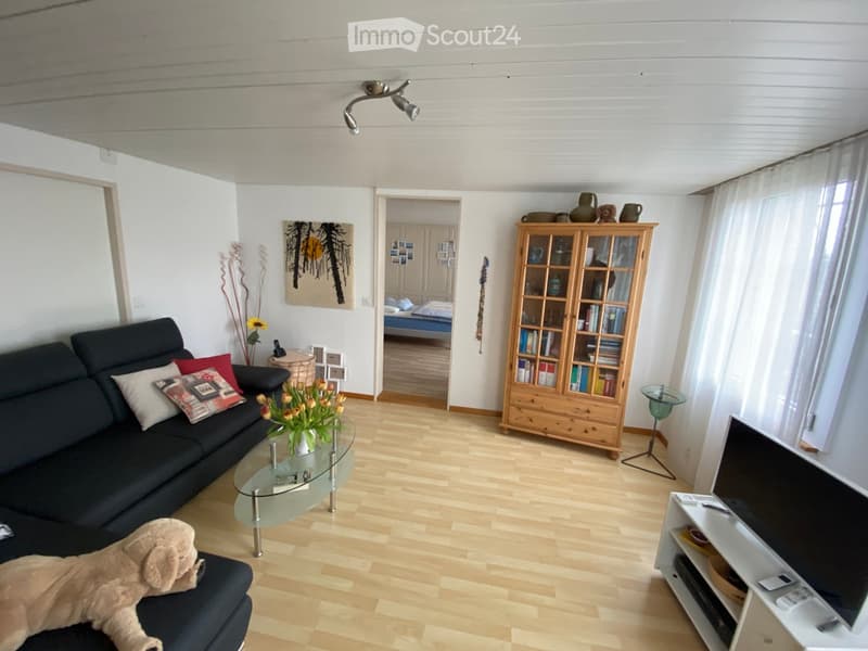 Bright living room with wooden flooring, a black sofa with decorative pillows, a glass coffee table, a wooden bookcase, a TV stand, and a view into a bedroom.