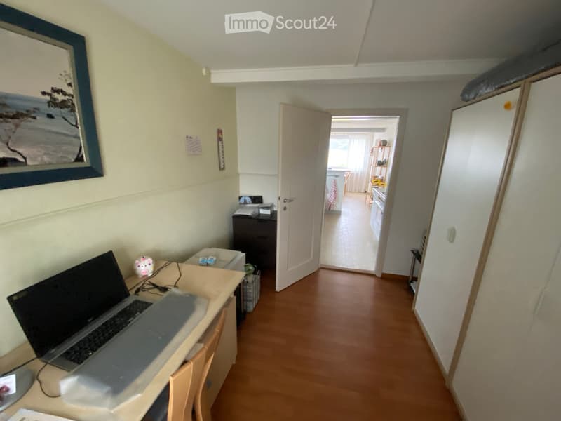 A furnished room with a wooden floor, a desk with a laptop, a chair, a cabinet, and a picture frame on the wall. An open door leads to another room with a bed and a nightstand.