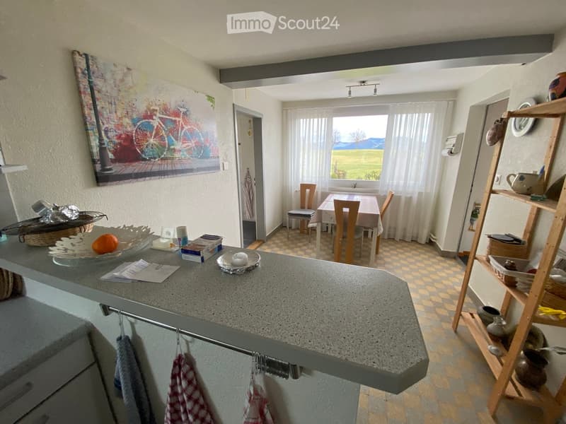 A modern kitchen with a grey countertop, a dining area with a table and chairs, and a shelf with decorative items. The room features a large window with a scenic view, a colorful painting on the wall, and a tiled floor.