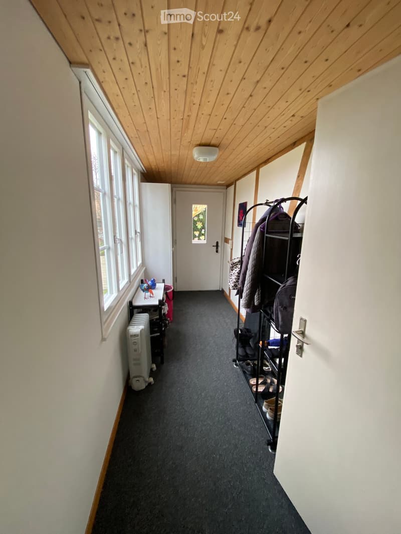 A narrow hallway with wooden slanted ceiling, carpeted floor, and white walls. Features include a coat rack with jackets and shoes, a small table with a decorative item, and a radiator on the left side.