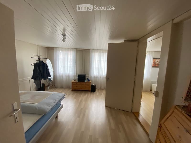 Bright bedroom with wooden flooring, a single bed, a wardrobe, a chest of drawers, and a mirror. The room has large windows with white curtains and an open doorway leading to another room.