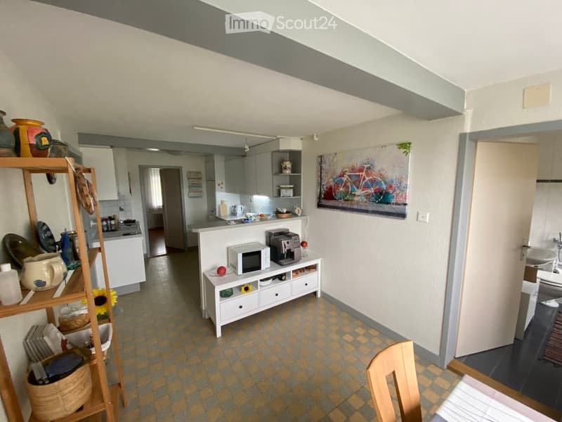 A cozy, well-lit kitchen and dining area with a checkered floor, white cabinets, a small table with a microwave and coffee maker, a wooden shelf with decorative items, and a painting on the wall.