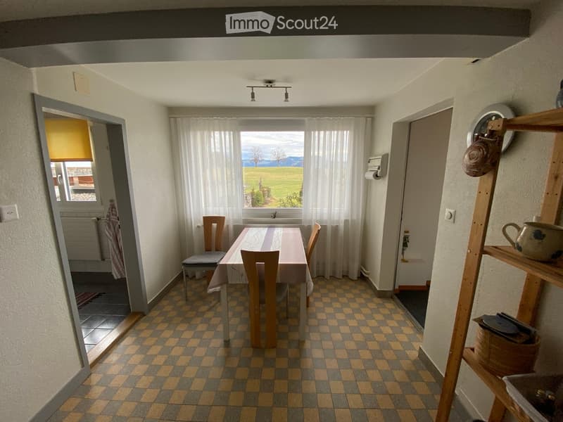 Bright kitchen with checkered floor, large window with white curtains, and a view of a green field. Includes a dining table with chairs and a wooden shelf with various items.