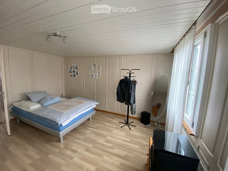 Bright bedroom with wooden flooring, a bed with blue bedding, a large mirror, a clothing rack with jackets, and picture frames on the wall.