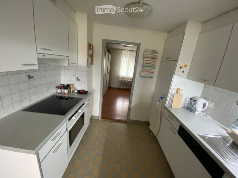 The kitchen features white cabinets, a tiled backsplash, and a grey countertop. Visible appliances include an oven, a sink, and a faucet. There are also various kitchen items such as a cutting board, bottles, and a kettle on the counter.