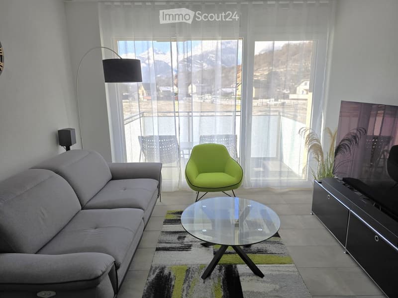 Modern living room with a grey sectional sofa, a round glass coffee table, a bright green armchair, a flat-screen TV on a black stand, and a floor lamp near large windows with sheer curtains.