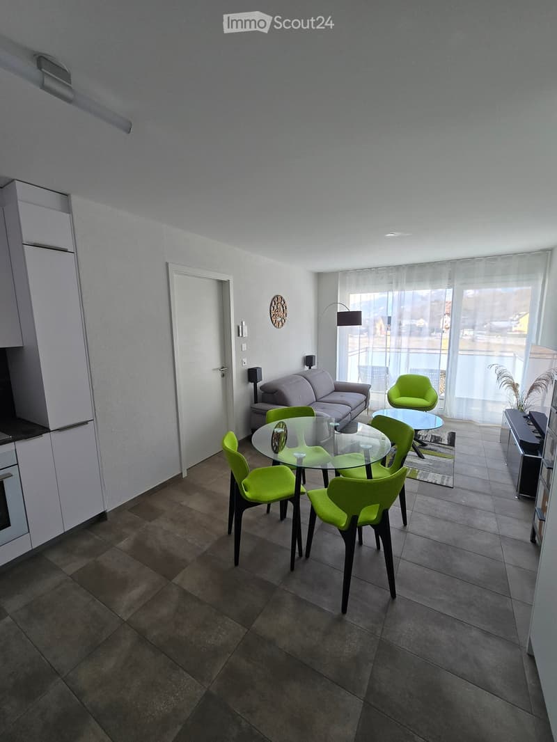Modern living and dining area with a grey sofa, glass dining table, green chairs, and a floor lamp near large windows.