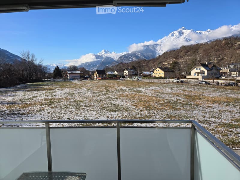 Balcony with a scenic view of snow-covered mountains and a town below, featuring a metal railing and a glass table.