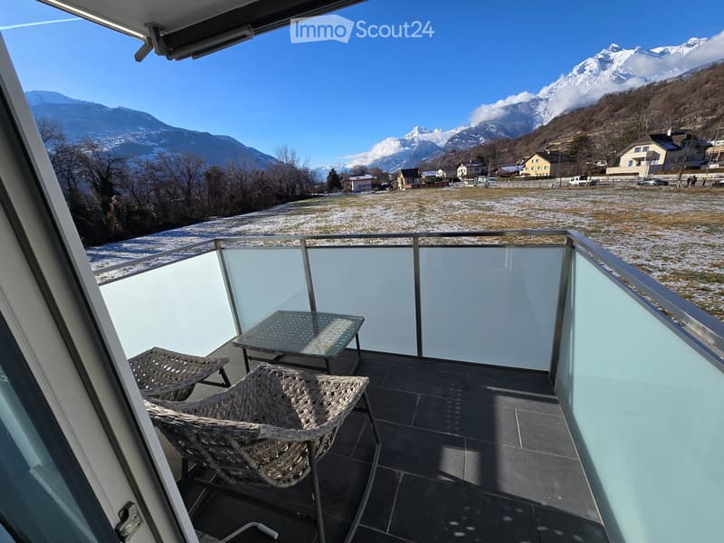 Balcony with glass railing, two wicker chairs, and a small table, overlooking a snowy landscape and mountains.
