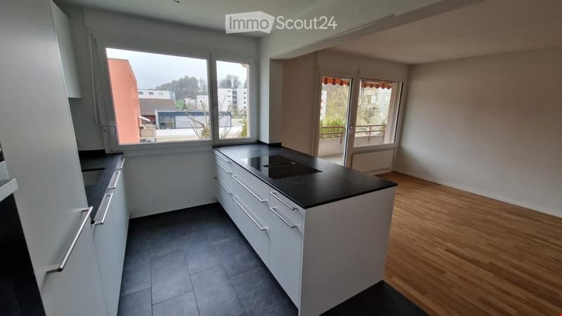 Modern kitchen with white cabinets, black countertop, and a central island. Large windows provide natural light, and the floor is wooden.