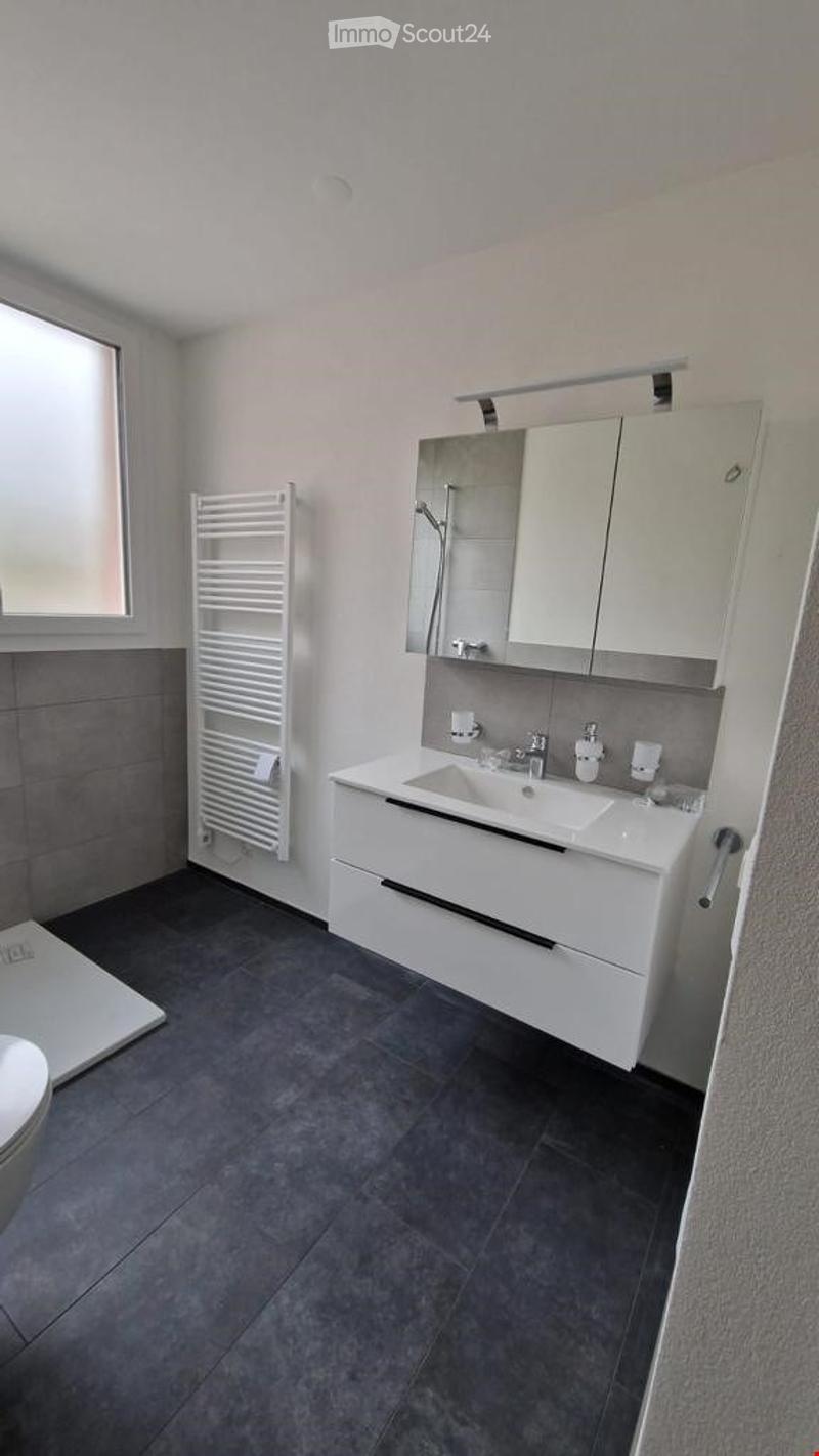 Modern bathroom with a white sink, mirrored cabinet, towel rack, and a toilet. The space features grey tiles on the floor and walls, and a window providing natural light.