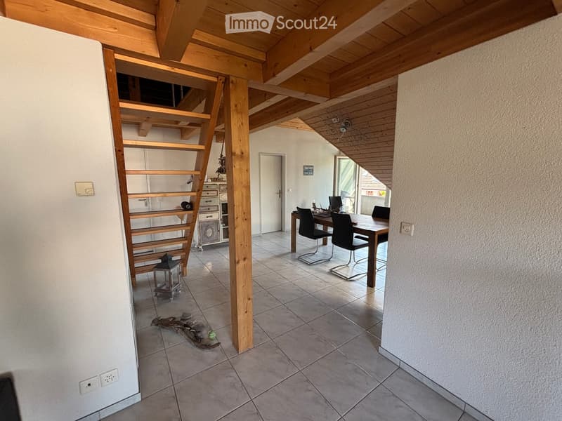 Bright, open room with wooden beams and tiled floor, featuring a dining table with chairs, a staircase leading to an upper level, and a small cabinet with drawers.