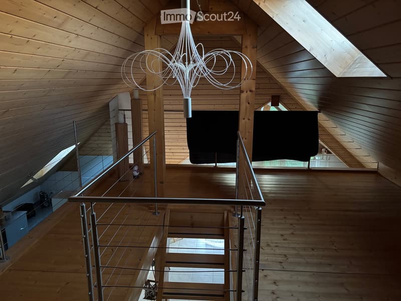 Open wooden hallway with a modern metal railing, exposed wooden beams, and a hanging light fixture. The space features large windows and a view of the lower level.