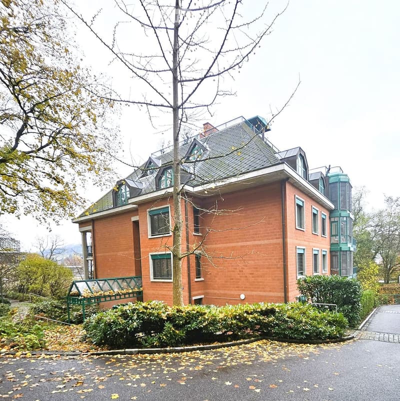 Two-story brick house with multiple windows, green shutters, and a sloped roof. Features a small balcony and surrounding greenery.