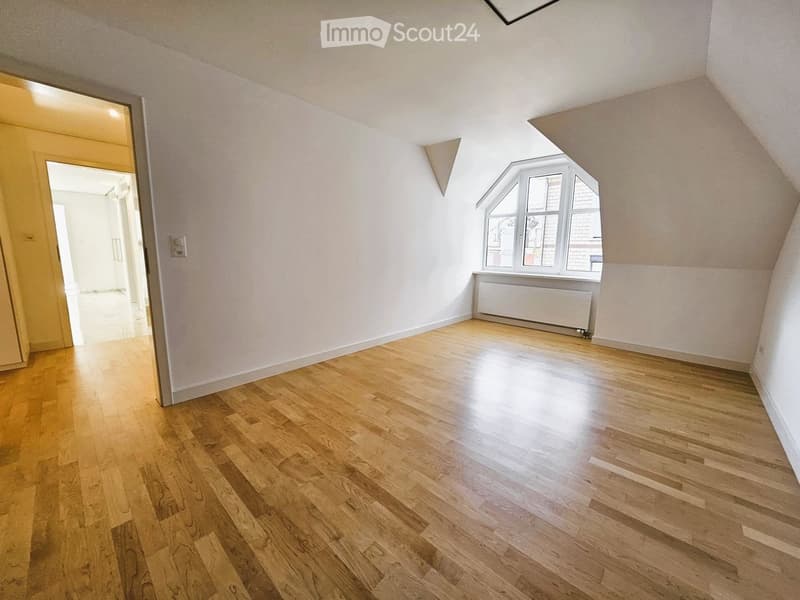 Empty attic room with wooden flooring, white walls, and a large window with a radiator below it. The room has a slanted ceiling and an open doorway leading to another area.