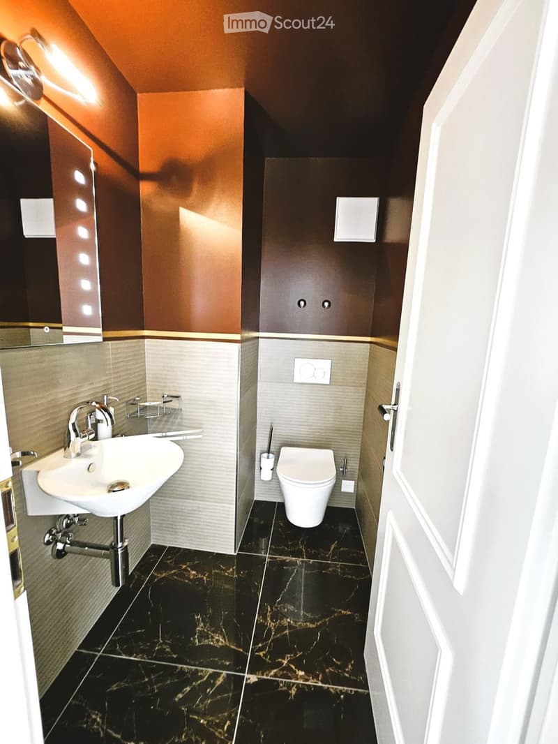 Modern bathroom with black marble flooring, white sink, chrome faucet, toilet, and a large mirror with built-in lighting.
