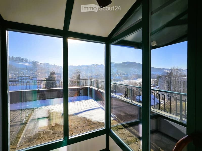 Spacious balcony with large glass windows offering panoramic views of a town and surrounding mountains, featuring tiled flooring and metal railings.