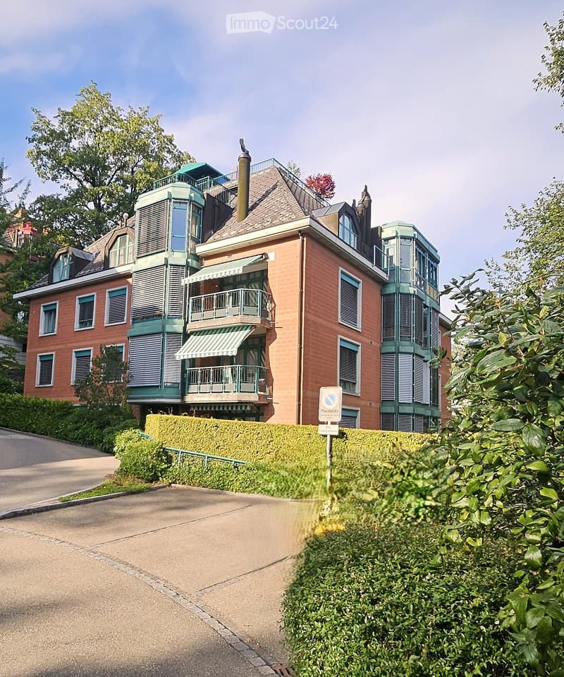 A multi-story brick house with green balconies, multiple windows, and a well-maintained hedge in front.