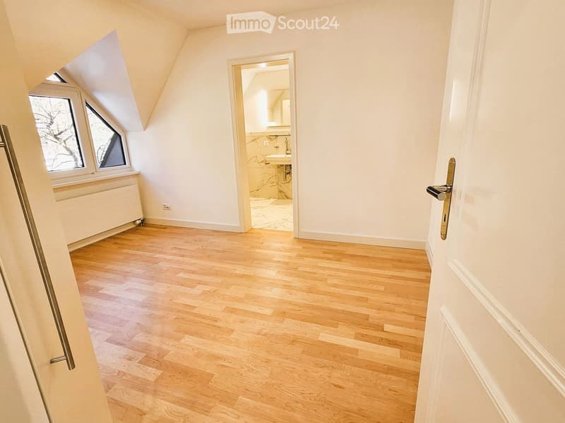 Empty room with wooden flooring, white walls, and a door leading to a bathroom. The room has slanted ceilings and two windows with a view of the outside.
