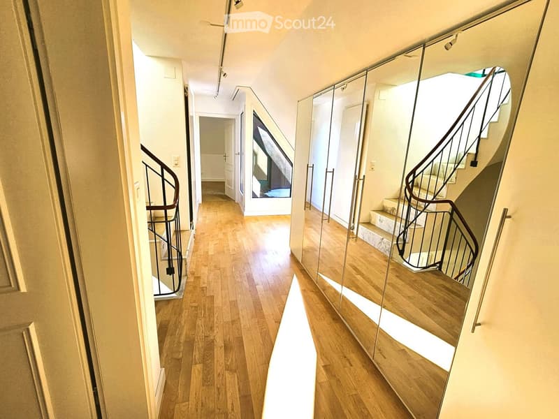 A modern hallway with wooden flooring, white walls, and two spiral staircases with black railings on either side. The hallway features large mirrors and recessed lighting.