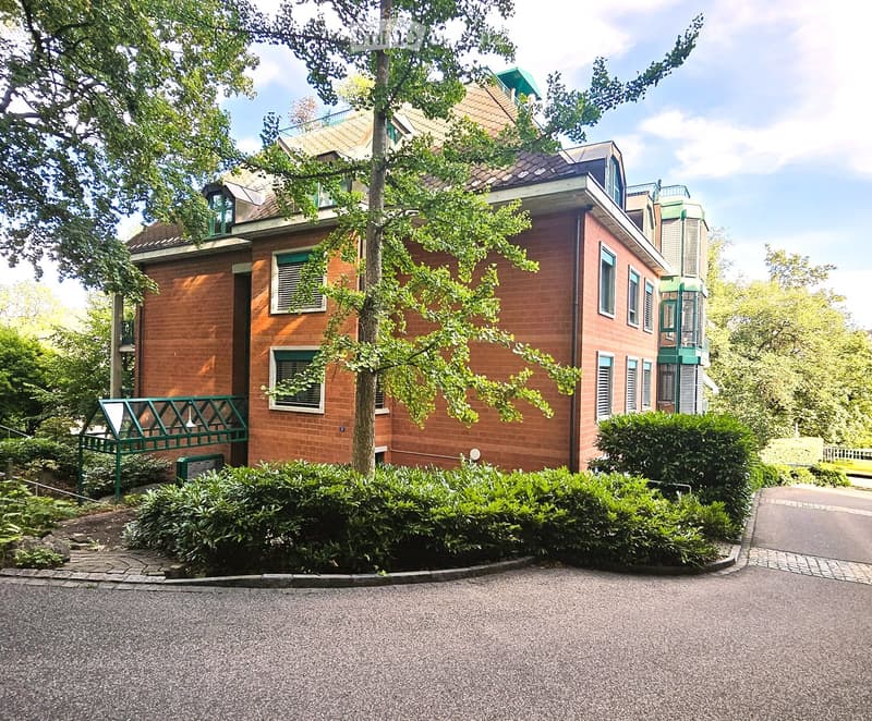 A two-story brick building with multiple windows, green shutters, and a small balcony. The building is surrounded by greenery and a paved driveway.