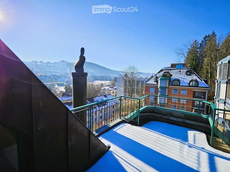 Balcony with green metal railing, overlooking a cityscape with buildings, trees, and distant mountains under a clear blue sky.