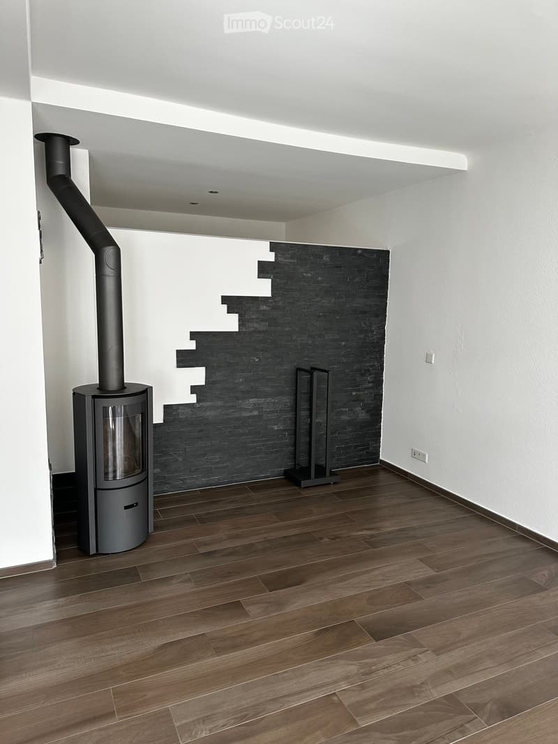 Modern unfurnished room with wooden flooring, white walls, and a black brick accent wall. Features a contemporary wood-burning stove with a tall chimney pipe.