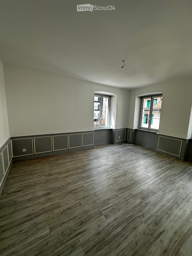 Empty room with wooden flooring, light green walls, and two windows allowing natural light to enter.