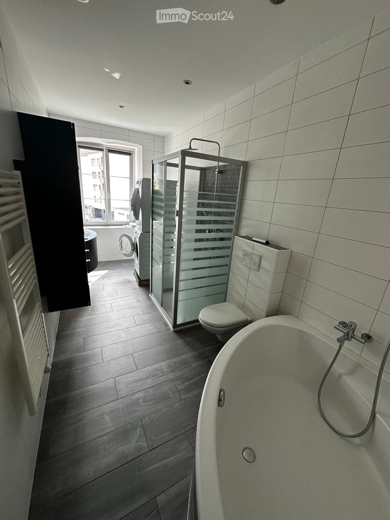 Modern bathroom with a bathtub, toilet, glass-enclosed shower, and radiator. The space features light grey tiles and a wooden floor.