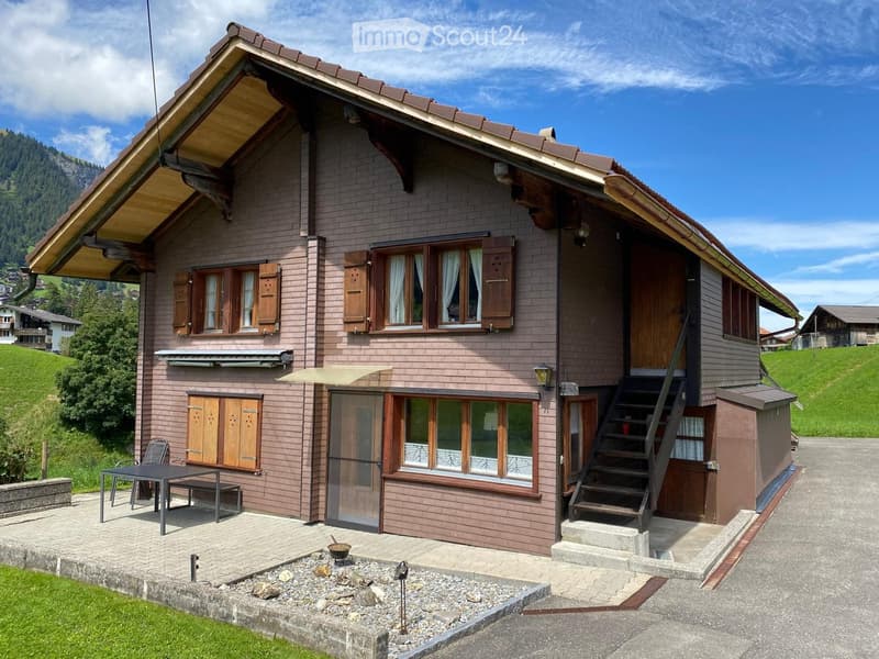 Two-story house with brown brick exterior, wooden shutters, and a raised ground floor entrance. Features include a patio with a table and chairs, a small fire pit, and a staircase leading to an upper level.