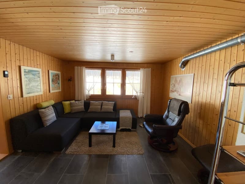Cozy living room with wooden walls and ceiling, dark grey sofa with multiple cushions, coffee table, armchair, and artwork on the walls.
