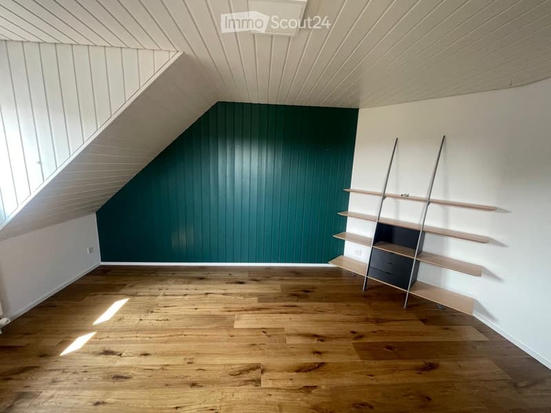 An empty attic room with wooden flooring, white walls, and a slanted ceiling. A single window allows natural light to enter, and a simple wooden shelving unit is mounted on one wall.