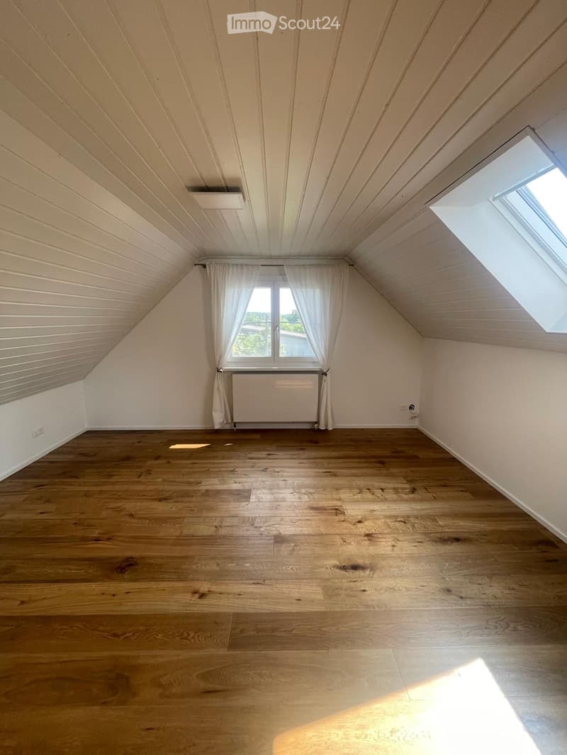 Empty attic room with wooden flooring, white walls, and a slanted ceiling. Features a window with white curtains and a radiator below it. The room has a skylight on the right side.
