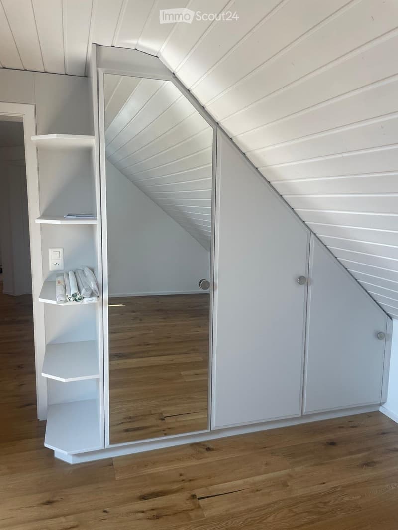 A modern, minimalist room with a large, full-length mirror, white built-in shelves, and neatly arranged towels. The room features wooden flooring and white walls.