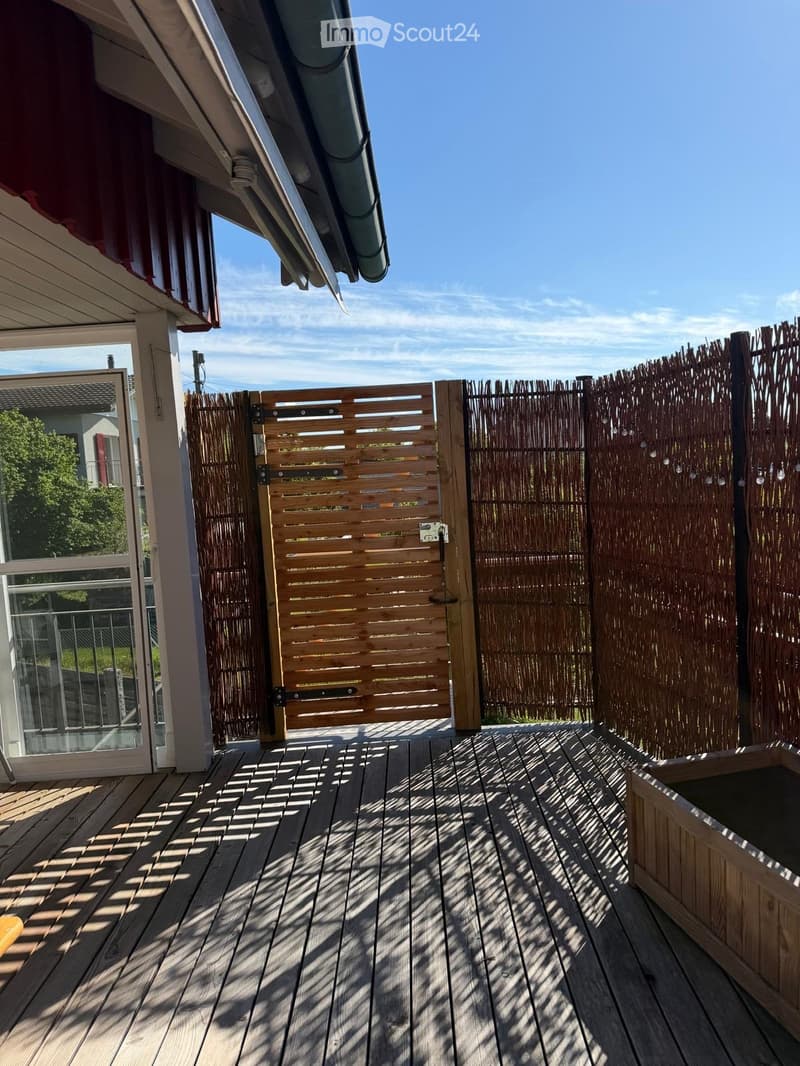 Wooden deck with a wooden fence and a glass door leading to the interior, with shadows cast by the fence on the deck.