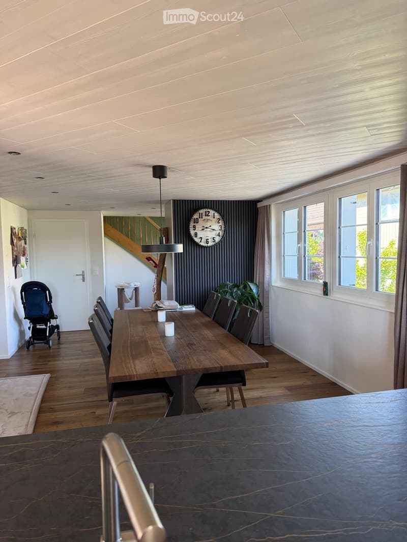 Dining room with wooden dining table, chairs, baby stroller, clock, and plant near the window.
