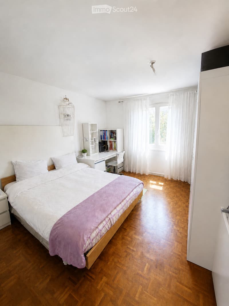 Bright bedroom with wooden herringbone flooring, a double bed with white bedding and a pink throw, a white desk with a chair, a small bookshelf, and a decorative wicker basket on the wall.