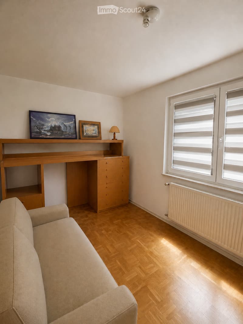 Living room with wooden flooring, a beige sofa, a wooden desk with drawers, a table lamp, and two picture frames on the desk.