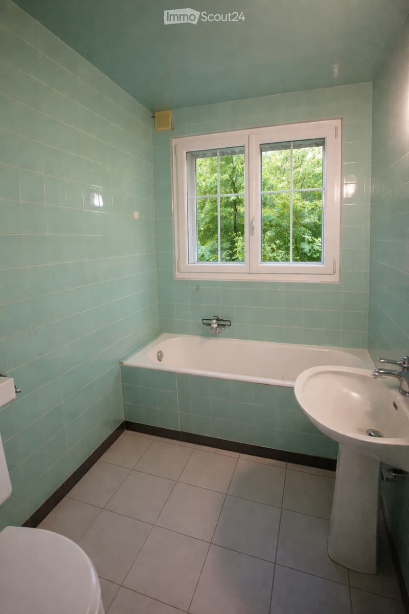 Bathroom with mint green tiled walls and ceiling, white bathtub, sink, and toilet, large window with a view of greenery.